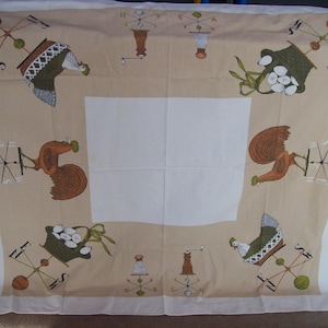 May include: A beige tablecloth with a white center square. The border features a repeating pattern of roosters, baskets of eggs, and wind vanes with the letters N, S, E, and W.