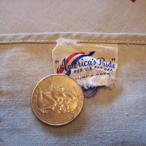 Pu&ograve; includere: Primo piano di un tessuto bianco con un'etichetta "America's Pride" cucita in rosso, bianco e blu. L'etichetta riporta il testo "Reg. U.S. Pat. Off." e una moneta da 25 centesimi &egrave; posizionata sul tessuto per la scala.