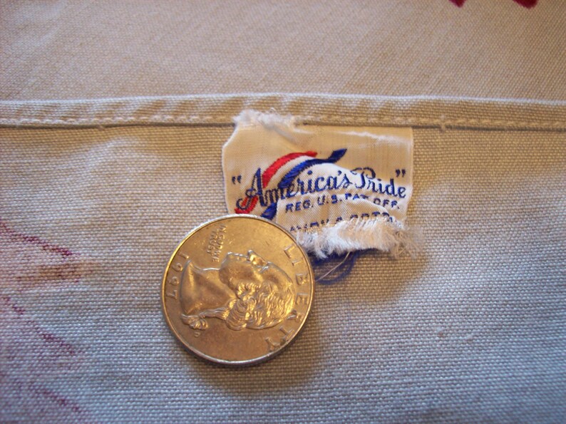 Pu&ograve; includere: Primo piano di un tessuto bianco con un'etichetta "America's Pride" cucita in rosso, bianco e blu. L'etichetta riporta il testo "Reg. U.S. Pat. Off." e una moneta da 25 centesimi &egrave; posizionata sul tessuto per la scala.