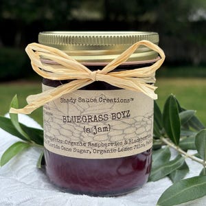 May include: A jar of "Bluegrass Boyz" jam with a gold lid and a raffia bow. The label lists ingredients like organic raspberries and blackberries. The jar sits on a white cloth with green foliage in the background.