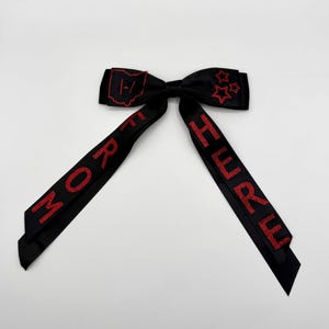 May include: A black hair bow featuring red glitter embroidery. The left side displays an outline of Ohio with a symbol inside, while the right side has three stars. The long tails feature the red glitter text FROM and HURU vertically.