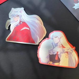 May include: Two anime-style stickers featuring characters with long white hair and detailed clothing. One sticker shows a character in a red outfit, while the other is in a cream-colored garment with a fur collar. Both stickers have a glossy finish.