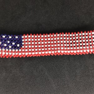 Puede incluir: Un brazalete de cuentas en los colores de la bandera estadounidense. El brazalete está hecho de cuentas rojas, blancas y azules y está dispuesto en un patrón que se asemeja a la bandera estadounidense.