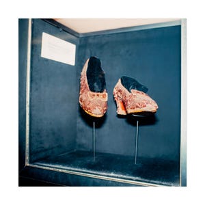 Può includere: Un paio di scarpe antiche e ornate in mostra. Le scarpe sono di colore marrone-rossastro con un motivo strutturato e piccoli tacchi. Sono esposte in una teca di vetro su uno sfondo blu scuro.