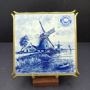 May include: A square ceramic tile featuring a blue and white hand-painted scene of a Dutch windmill, a house, and a small boat on water. The tile is encased in a gold-colored metal frame and includes a circular sticker that reads Delft Blue.