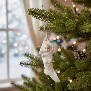 May include: A white ceramic Christmas ornament shaped like a stocking, with a baby figure inside. The baby is wearing a blue shirt and the stocking reads "Baby's First Christmas 1994". The ornament hangs from a silver string on a Christmas tree.