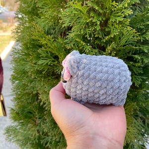 May include: A grey crocheted sheep plush toy with pink ears and a black nose. The toy is being held in a person's hand.