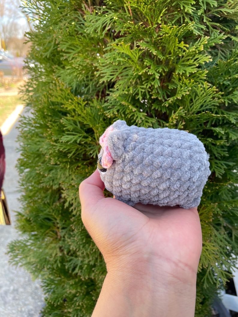 May include: A grey crocheted sheep plush toy with pink ears and a black nose. The toy is being held in a person's hand.