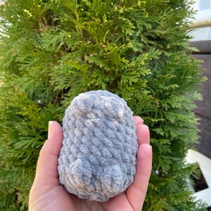 May include: A grey crocheted stuffed animal in the shape of a penguin. The penguin has a textured surface and is being held in a person's hand.