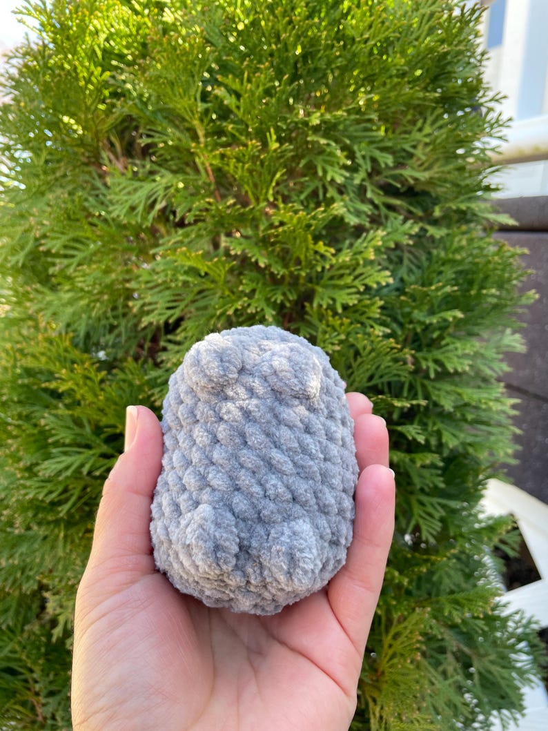 May include: A grey crocheted stuffed animal in the shape of a penguin. The penguin has a textured surface and is being held in a person's hand.