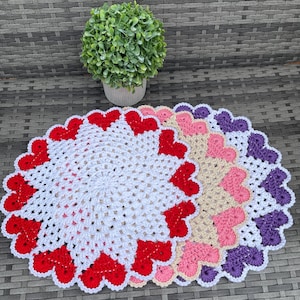 May include: A collection of handmade crochet doilies in various colors, including white with red hearts, beige with pink hearts, and white with purple hearts. The doilies are arranged on a gray surface, with a small potted plant in the background.