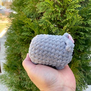 May include: A grey crocheted sheep plush toy with a pink bow. The toy is being held in a person's hand.