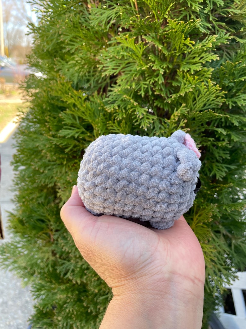 May include: A grey crocheted sheep plush toy with a pink bow. The toy is being held in a person's hand.