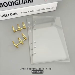 May include: Two clear rectangular plastic sheets with pre-punched holes along one side and two separate gold-colored metal three-ring binder mechanisms. The text 2pcs board+2 gold ring 6 Flavors is printed at the bottom of the image.