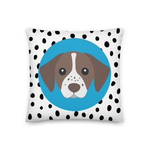 May include: A white pillow with black polka dots and a blue circle in the center. Inside the circle is a brown and white dog with brown ears and a black nose.