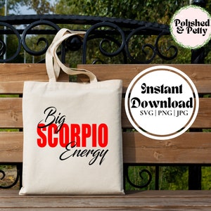 May include: A white tote bag with black text that says "Big Scorpio Energy" in red letters. The bag is sitting on a wooden bench.