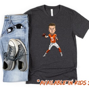 May include: A dark gray t-shirt with a cartoon illustration of a football player in an orange and white uniform. The text "AVAILABLE IN KIDS SIZES!" is printed on the bottom of the shirt.