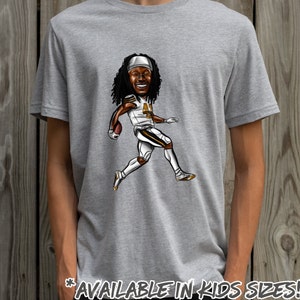 May include: A gray t-shirt with a cartoon illustration of a football player running with the ball. The player is wearing a white jersey with gold trim and black pants. The text "AVAILABLE IN KIDS SIZES!" is at the bottom of the shirt.