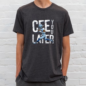 May include: Dark gray t-shirt with the text "CEE YOU LATER" in distressed white lettering. A football player in a blue and white uniform is running on the shirt. The shirt is being worn by a person.