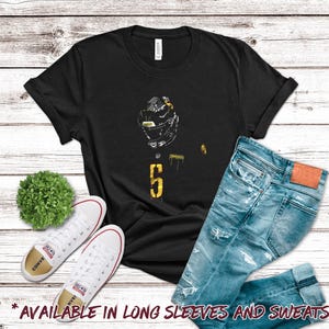 May include: Black t-shirt with a football helmet graphic and the number 5 in yellow. The shirt is paired with blue jeans and white sneakers. Text at the bottom says "AVAILABLE IN LONG SLEEVES AND SWEATSHIRTS!"