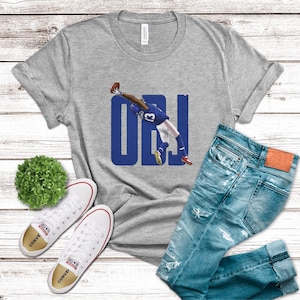 May include: A heather gray short-sleeve t-shirt featuring a graphic of a football player in a blue and white uniform making a one-handed catch, overlaid with the large blue text OBJ. The shirt is paired with blue distressed denim jeans.