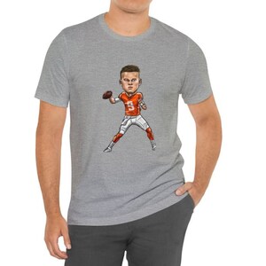 May include: A gray t-shirt with a cartoon illustration of a football player wearing an orange and white jersey and holding a football. The player is wearing a white helmet with a black face mask.