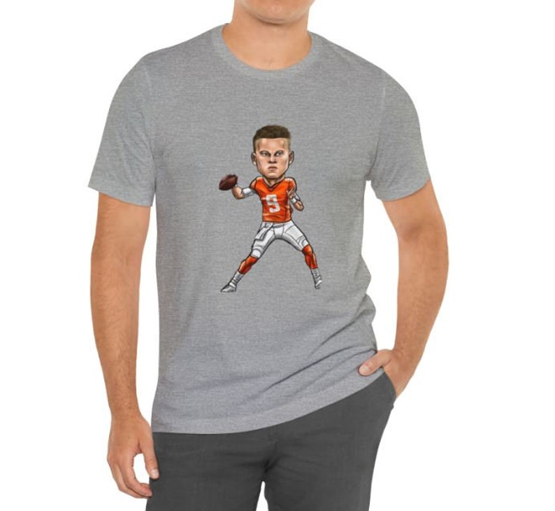 May include: A gray t-shirt with a cartoon illustration of a football player wearing an orange and white jersey and holding a football. The player is wearing a white helmet with a black face mask.