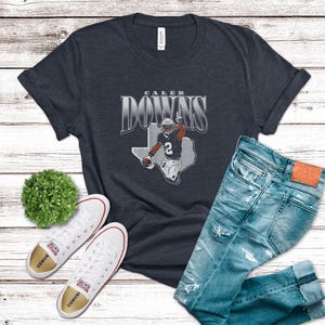 May include: A navy blue t-shirt with the text "Caleb Downs" above an illustration of a football player in a Texas outline. The shirt is paired with light blue jeans and white sneakers with red accents.