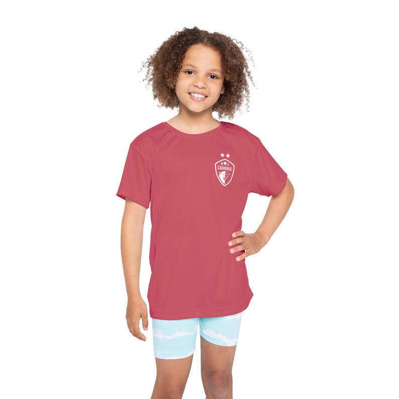 May include: A young person wearing a red short-sleeved t-shirt with a white graphic of a shield with the text "I'M COURAGE" and two stars. The person is also wearing light blue and white patterned shorts.