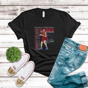 Op de afbeelding: Zwart T-shirt met een afbeelding van een basketballer in rood en wit, met de tekst "Atlanta Basketball" en "Brittney Griner" in wit. De speler houdt een basketbal vast en heeft een handtekening onder de afbeelding.