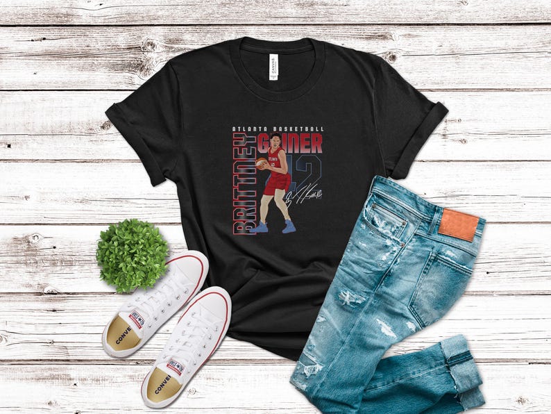 Op de afbeelding: Zwart T-shirt met een afbeelding van een basketballer in rood en wit, met de tekst "Atlanta Basketball" en "Brittney Griner" in wit. De speler houdt een basketbal vast en heeft een handtekening onder de afbeelding.
