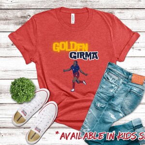May include: Red t-shirt with a yellow and blue graphic of a soccer player with the text "Golden Girma".