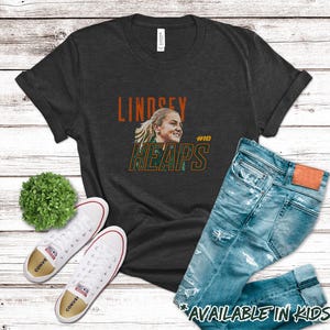 May include: Dark gray t-shirt with the name "Lindsey Heaps" and a photo of a woman. The shirt is paired with blue jeans and white sneakers. The text "AVAILABLE IN KIDS SIZES!" is at the bottom.