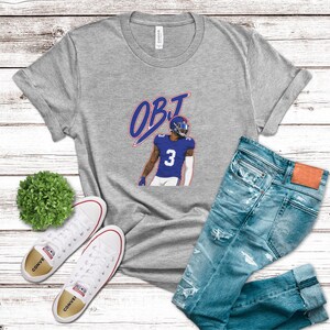 May include: A heather gray t-shirt featuring a graphic of a football player wearing a blue jersey with the number 3 and the text OBJ in blue and red. The shirt is displayed alongside a pair of distressed blue jeans and white canvas sneakers.