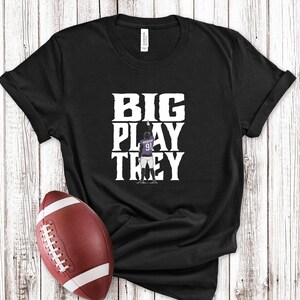May include: Black t-shirt with the text "BIG PLAY TREY" in white block letters. A football is in the foreground. The shirt features a football player in a purple jersey with the number 91.
