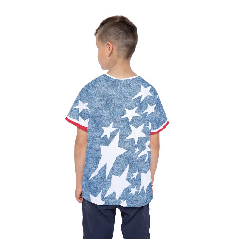 Puede incluir: Una camiseta de manga corta con un fondo azul efecto denim adornado con estrellas blancas de diferentes tama&ntilde;os. Las mangas tienen ribetes rojos y blancos. La camiseta la lleva una persona de espaldas, con pantalones azul marino.