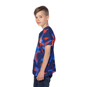 May include: A navy blue t-shirt with a red and blue geometric pattern. The shirt features a small emblem on the chest with the letters "USA". The shirt is short-sleeved.