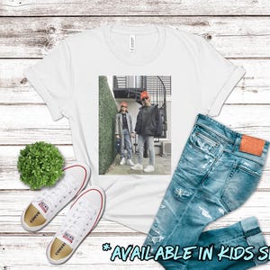 May include: White t-shirt with a graphic of two people wearing red hats and standing in front of a brick building with green plants. The text "AVAILABLE IN KIDS SIZES!" is at the bottom of the image.