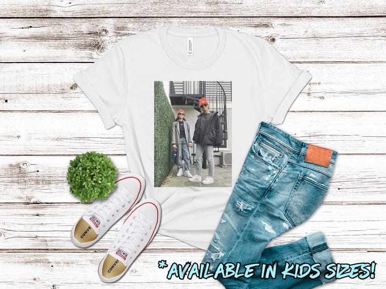 May include: White t-shirt with a graphic of two people wearing red hats and standing in front of a brick building with green plants. The text "AVAILABLE IN KIDS SIZES!" is at the bottom of the image.