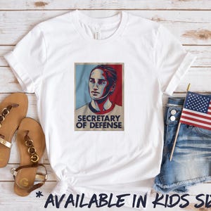May include: White t-shirt with a graphic of a woman's face and the text "Secretary of Defense".