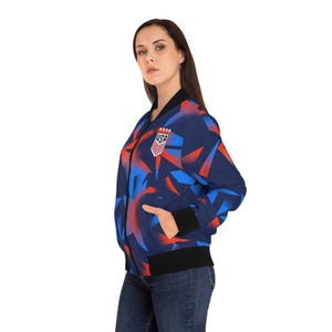 May include: A navy blue bomber jacket with a geometric pattern in red and blue. The jacket features a black collar, cuffs, and hem, and a USA emblem on the chest. The jacket is worn with blue jeans.