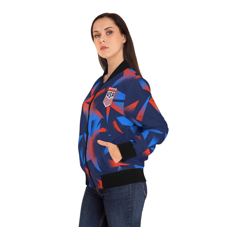 May include: A navy blue bomber jacket with a geometric pattern in red and blue. The jacket features a black collar, cuffs, and hem, and a USA emblem on the chest. The jacket is worn with blue jeans.