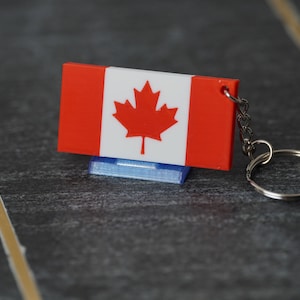 May include: A red and white Canadian flag keychain with a red maple leaf on a white background. The keychain is attached to a silver metal ring.