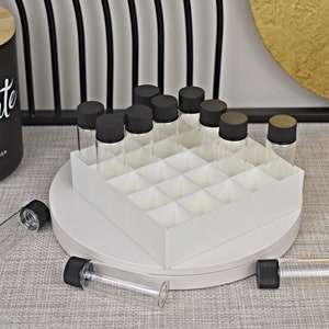 May include: A white organizer with 16 compartments for holding small glass bottles with black lids. The organizer is on a white circular base. There are several bottles with black lids on the surface next to the organizer.