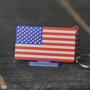 May include: A red, white and blue American flag keyring. The flag is made of plastic and has a silver metal key ring.