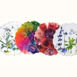 Può includere: Quattro sottobicchieri in resina con bordi organici e irregolari. Ogni sottobicchiere presenta un design unico: uno con fiori pressati, uno con un arcobaleno di colori, uno con tonalità rosso scuro e viola e uno con fiori blu. I sottobicchieri sono su sfondo bianco.