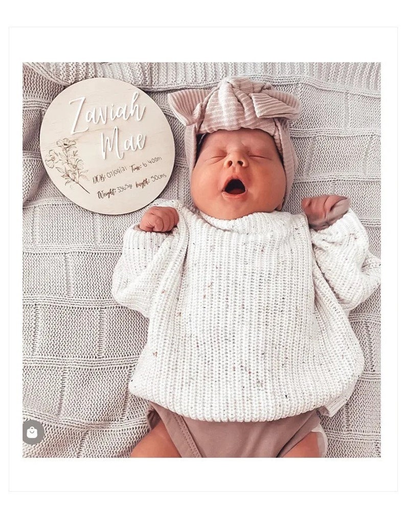 May include: A wooden baby announcement plaque with the name "Zariah Mae" and the date "01/01/21" along with the time "Time: 6:40am", weight "Weight: 3.5kg" and length "Length: 50cm". A baby girl wearing a white knitted sweater and a pink bow headband is lying on a gray blanket.