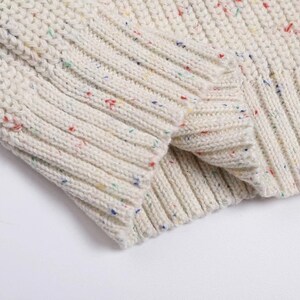May include: Close-up of a white knitted sweater with colorful flecks throughout the fabric. The sweater has a ribbed texture and a soft, cozy look.