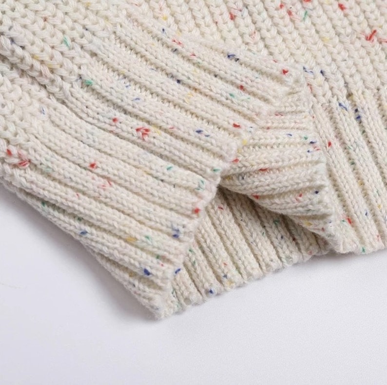 May include: Close-up of a white knitted sweater with colorful flecks throughout the fabric. The sweater has a ribbed texture and a soft, cozy look.
