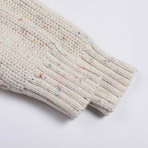 May include: Close-up of the cuff of a white knitted sweater with colorful flecks throughout the fabric.